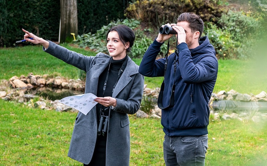 Twee mensen ontmoeten elkaar om vogels te kijken. De man kijkt door een verrekijker, terwijl de vrouw naar iets wijst.